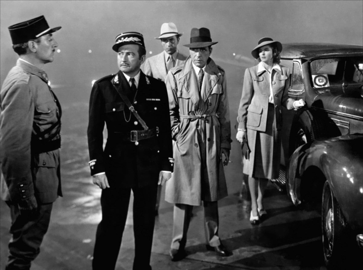 Humphrey Bogart wears a trench coat as Rick Blaine with Ingrid Bergman as Ilsa Lund, Claude Rains as Captain Louis Renault, and Paul Henreid as Victor Laszlo in the 1942 American romantic drama film Casablanca